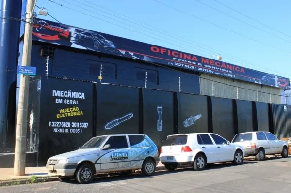 Lateral do galpão preto da oficina vista da rua, com o letreiro Mecânica em geral e injeção eletrônica e carros estacionados ao longo da calçada