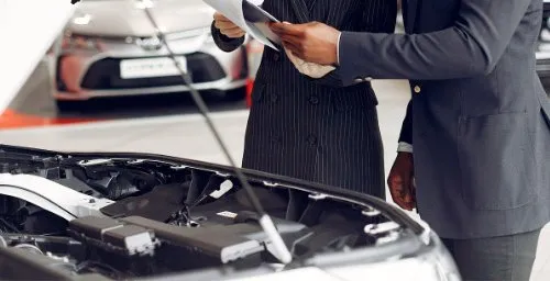 Atendente com prancheta anotando a revisão em frente a um carro com o capô aberto