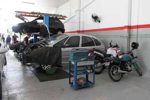 Salão de serviço com um carro no elevador ao fundo, outro coberto por capa de proteção e duas motos encostadas na parede